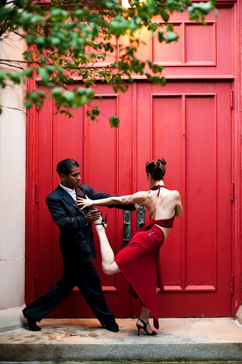 A man in a black suit and a woman in a red dress dance the Tango together.